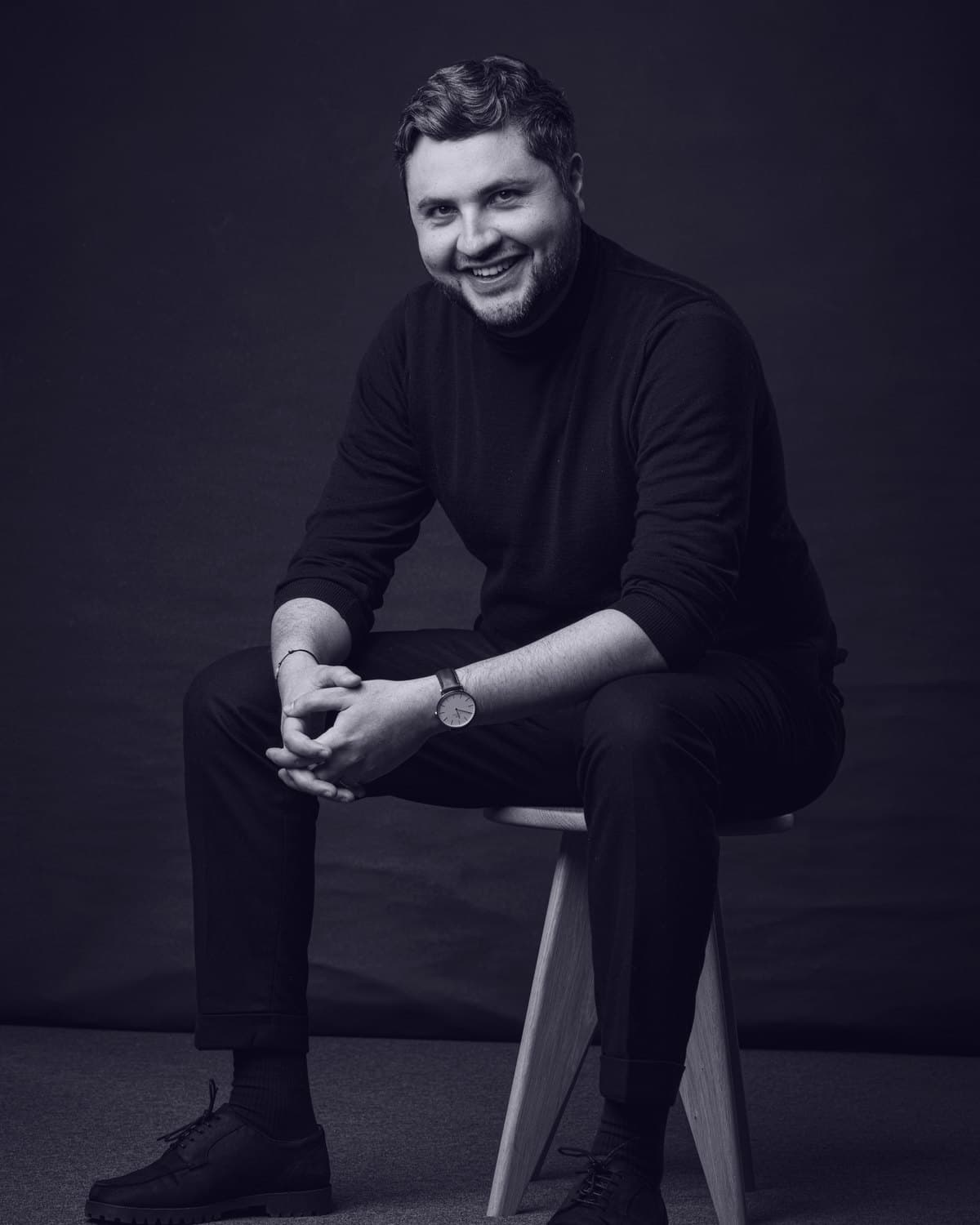 Karim Sandid, seated on a wooden stool in a black roll-neck, leaning forward and smiling